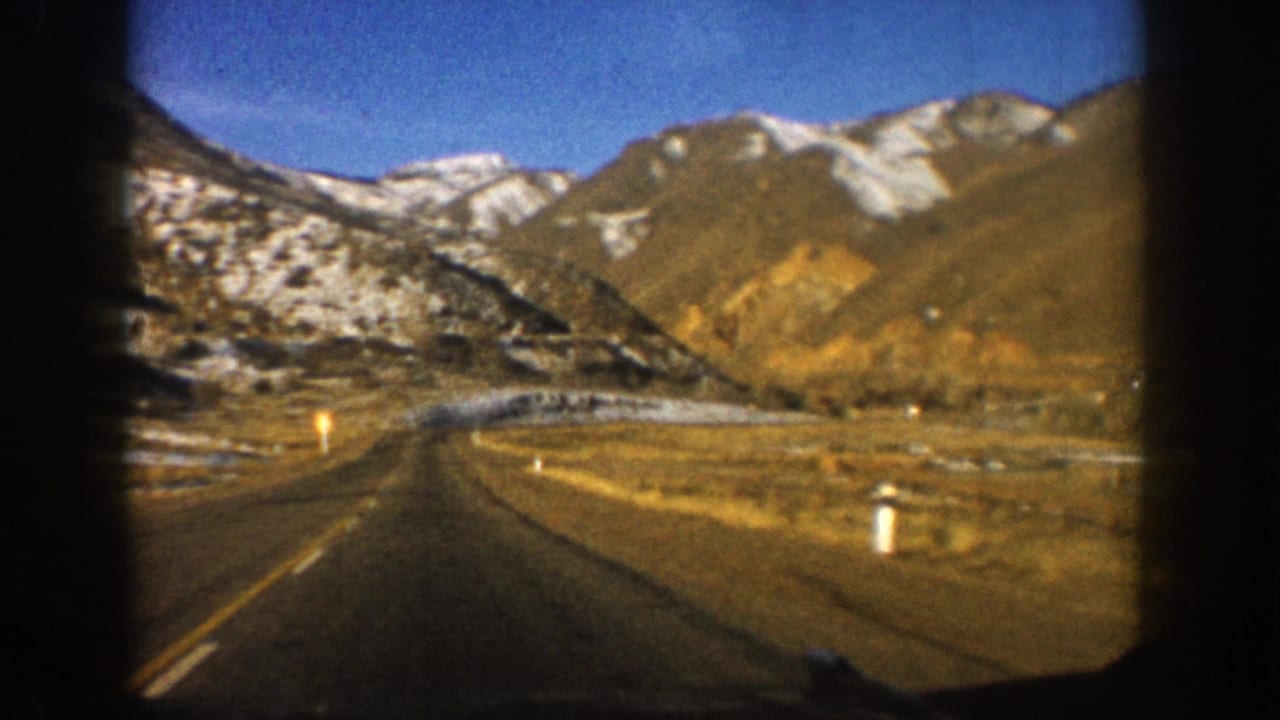8mm home-movie windshield view on a Colorado mountain highway, circa 1961, showing snow-capped peaks, arid hillside, and roadside delineator posts.