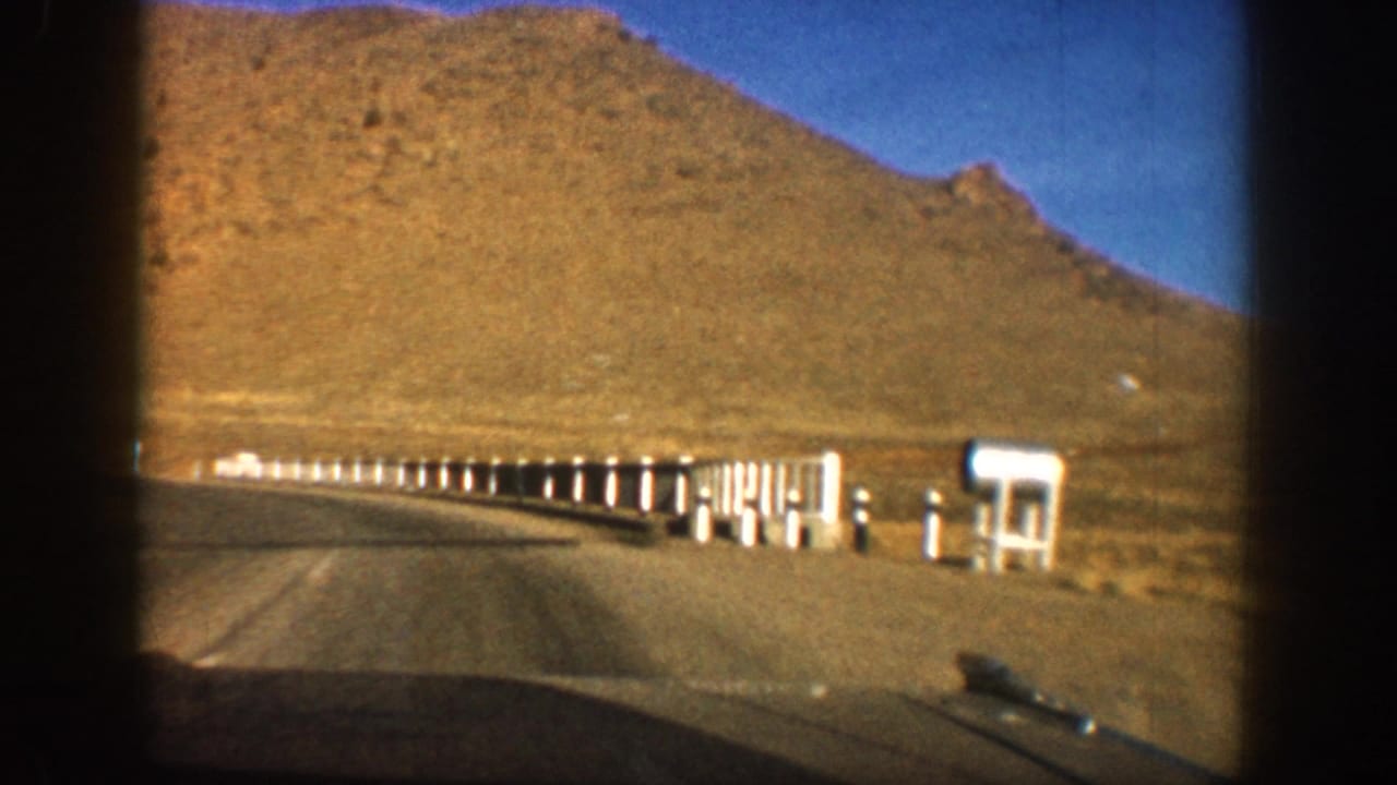 Windshield driving shot of a road curving right past white delineator posts and a large white roadside structure, with a brown hillside above.