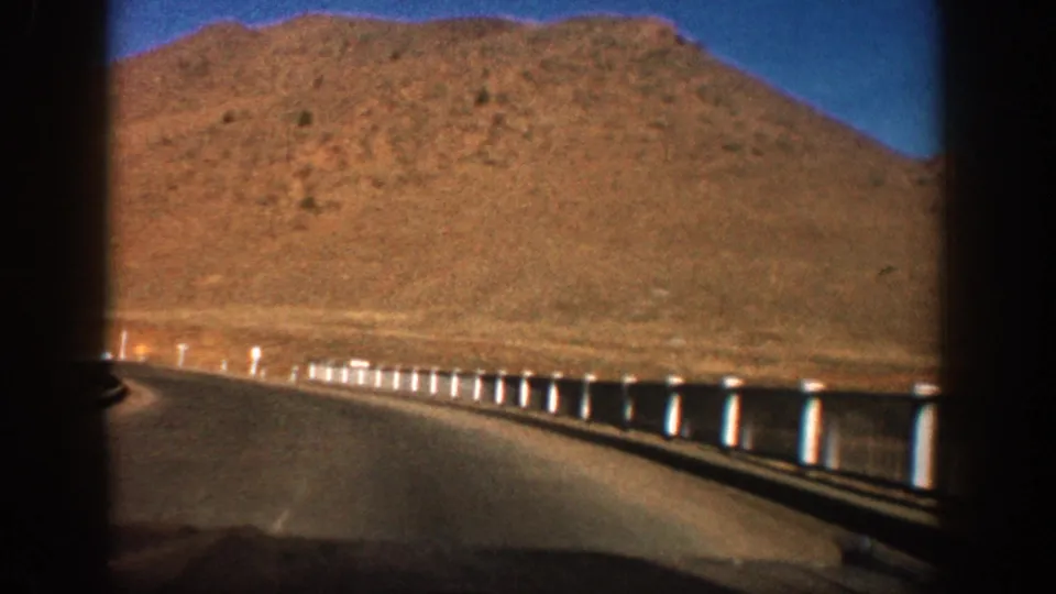 8mm home-movie driving shot on a Colorado mountain road, circa 1961, showing a pipe guardrail with white posts, arid red hillside, and distant snow-capped peaks.