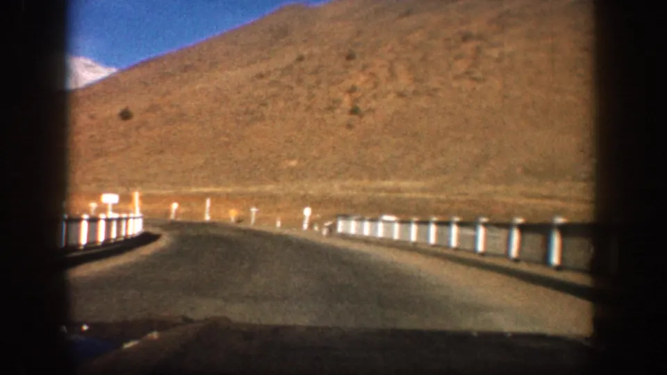 8mm film driving shot on a Colorado mountain highway, circa 1961, road curving past white guardrail posts with a barren red hillside under clear blue sky.