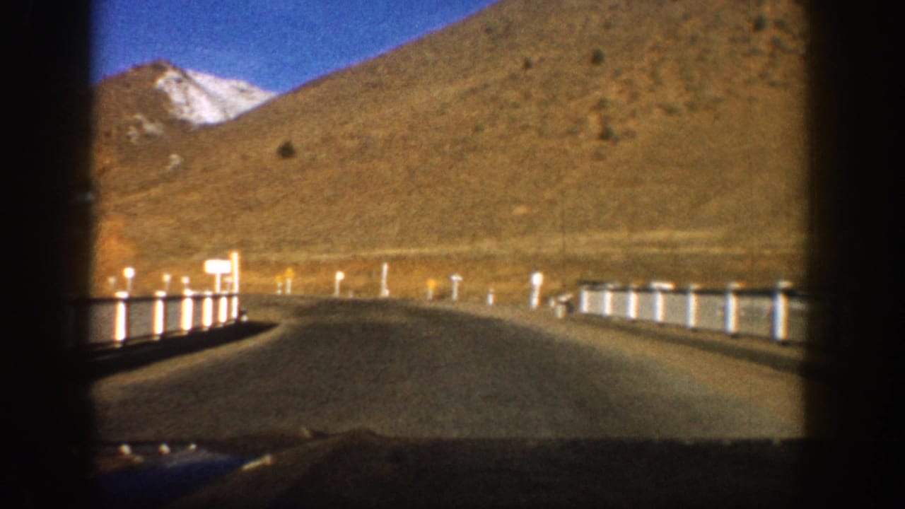 8mm film frame, c. 1961, looking down a two-lane mountain road in the Colorado Rockies, white delineator posts lining both sides, arid brown slopes and snow-capped peak under deep blue sky.