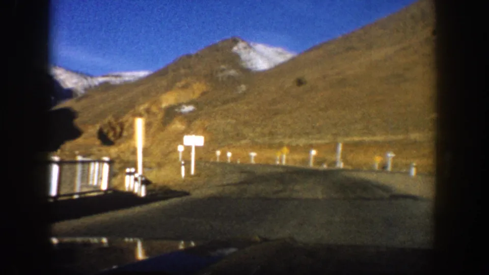 8mm home movie frame, c. 1961 Colorado, road view with white delineator posts, an unreadable roadside sign, brown mountain slope, and partial snow-covered summit under blue sky.