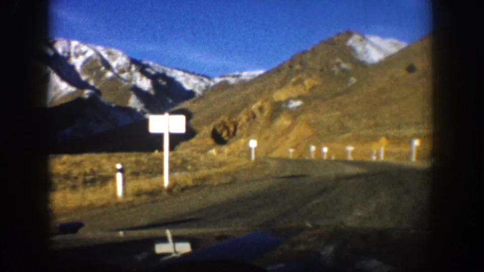 8mm film frame, c. 1961 Colorado, mountain road with white rectangular road sign at left, delineator markers, snow-capped peaks rising behind brown valley floor under blue sky.