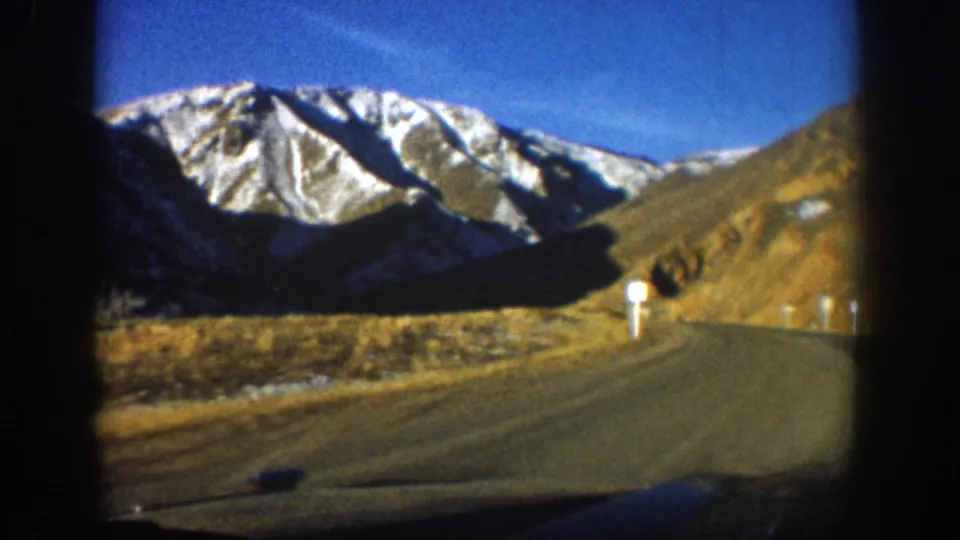 8mm film frame, c. 1961 Colorado Rockies — dramatic snow-covered peaks dominate the upper frame, brown rocky slopes below, road with white delineator posts at bottom, deep blue sky.