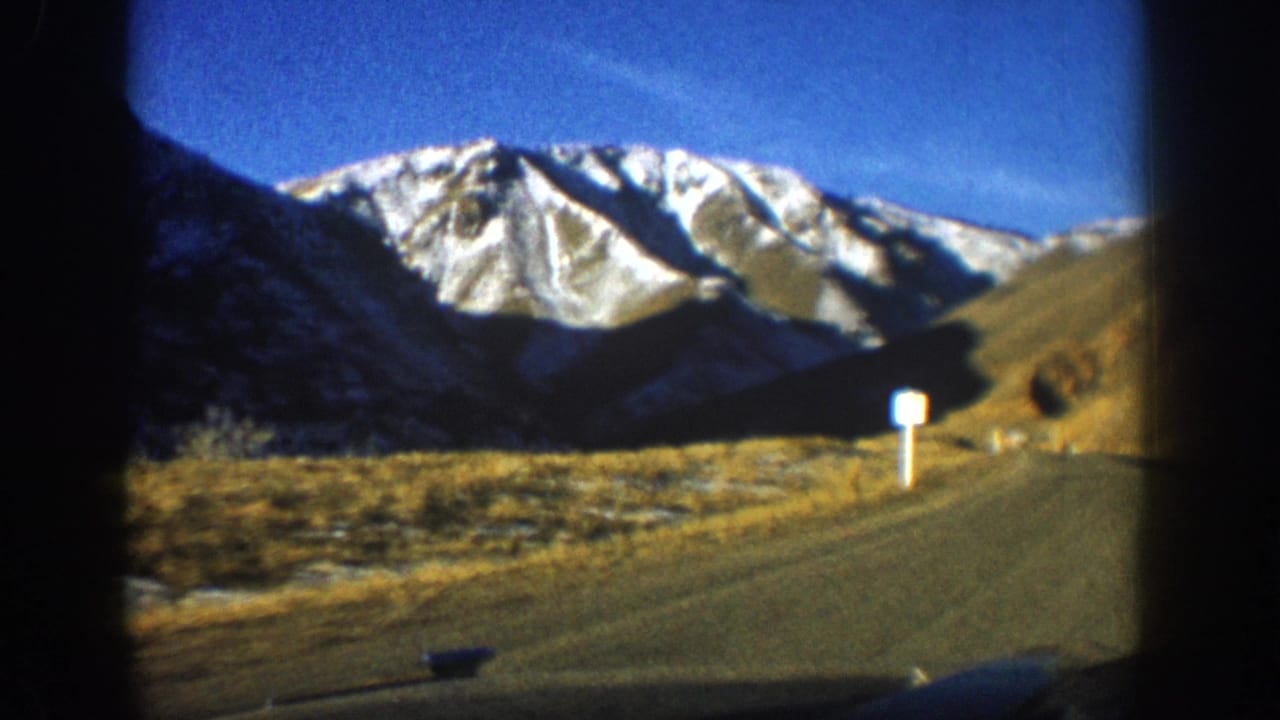 8mm home movie frame, circa 1961, Aspen area Colorado — snow-capped Rockies seen through windshield, golden autumn grasses, paved road, white marker post.