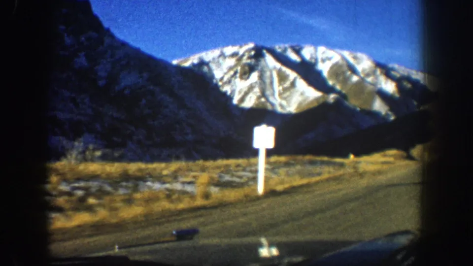 8mm home movie, circa 1961 Aspen area — snow-capped peaks, autumn hillside, white rectangular road sign on post visible, with right-edge obstruction in frame.