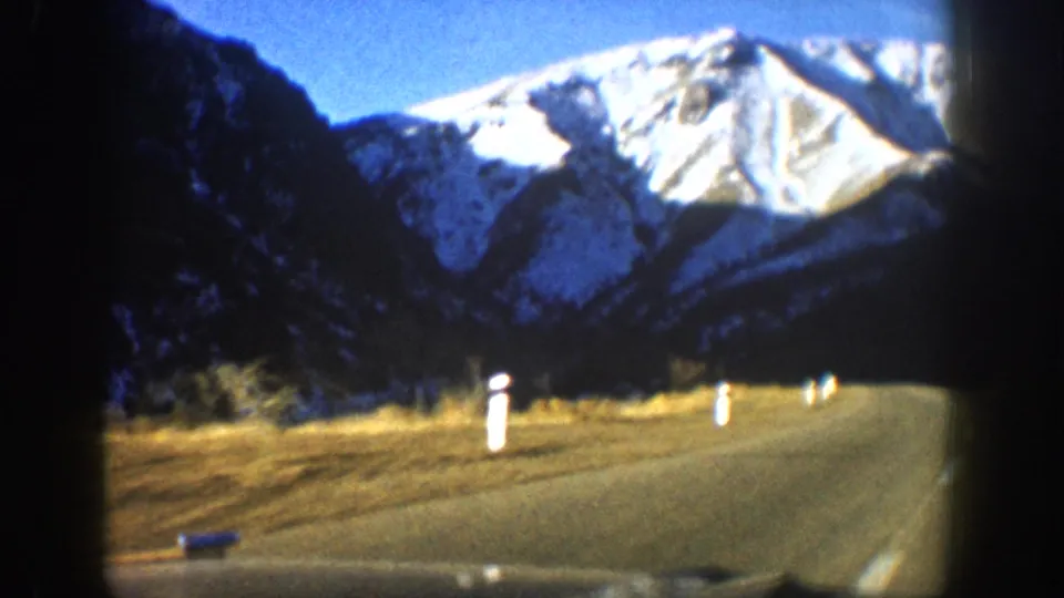 8mm home movie, circa 1961 Aspen Colorado area — closer angle on snow-capped ridge, dry autumn grasses, multiple white road markers along highway edge.