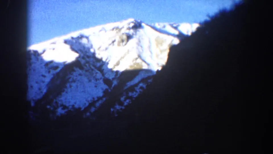 8mm home movie, circa 1961 Aspen Colorado area — dramatic close angle on snow-covered Rocky Mountain peak with dark rocky or structural element at right edge.