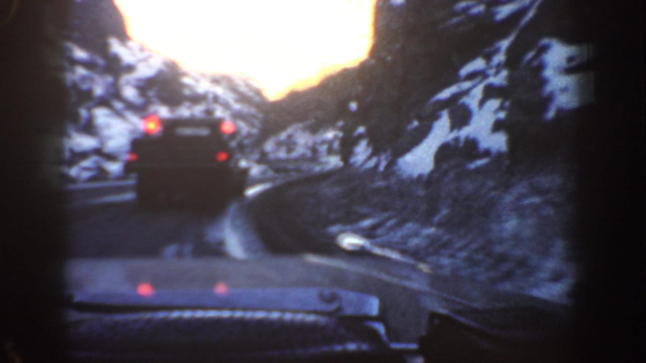 Dashboard POV of a dark vehicle ahead with red brake lights on a winding mountain road flanked by snow-covered slopes.