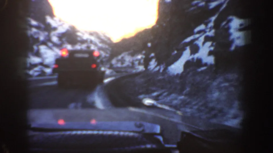 Windshield POV following a dark vehicle with red brake lights on a narrow winding mountain road, flanked by snow-dusted rocky slopes, shot on 8mm film circa 1961 in the Colorado Rockies.