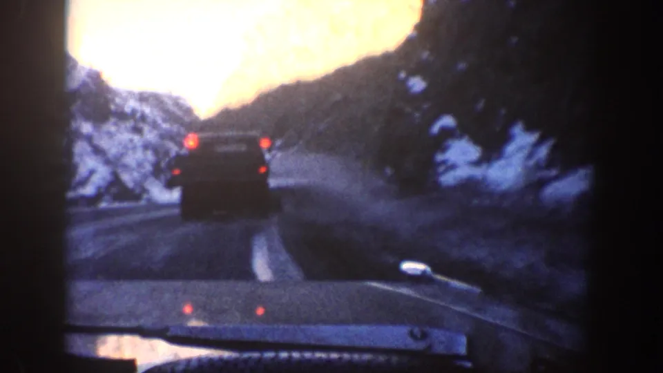 Interior windshield shot following a dark vehicle on a winding mountain road, snow-covered slopes on both sides, overexposed orange sky — 8mm home-movie footage from a 1961 Colorado mountain drive.