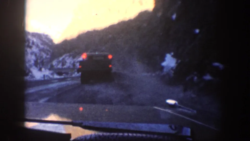 Windshield POV of a dark vehicle receding ahead on a winding mountain road, snow-covered slopes framing both sides — closing frames of a 1961 8mm Colorado mountain road sequence.