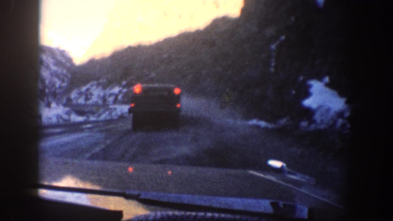 8mm home-movie shot through a windshield following a 1960s-era car on a snow-lined mountain road, likely in Colorado circa 1961, with a warm orange light leak at upper left.