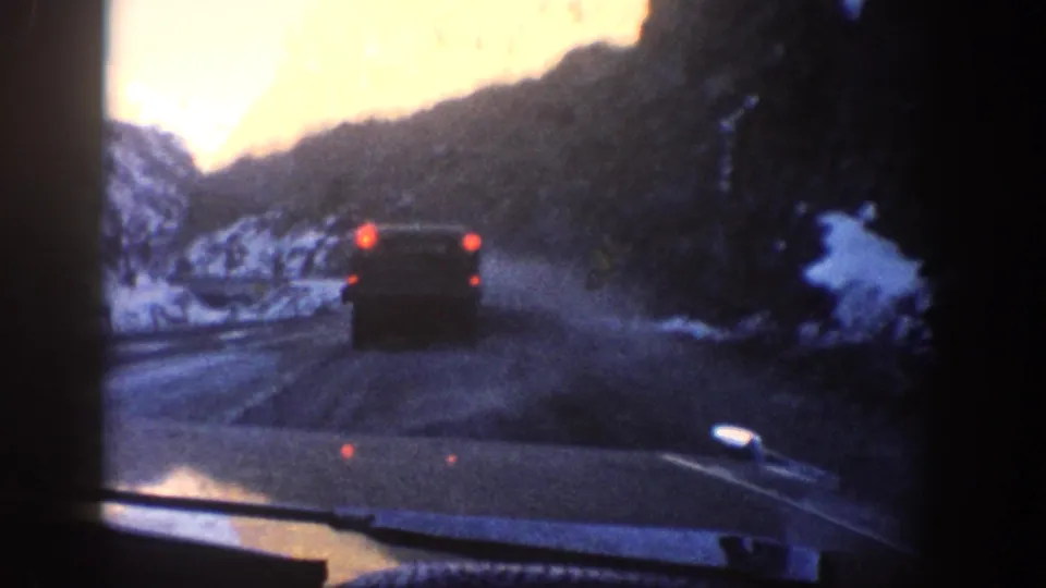 8mm home-movie shot through a windshield following a 1960s-era car on a snow-lined mountain road, likely in Colorado circa 1961, with a warm orange light leak at upper left.