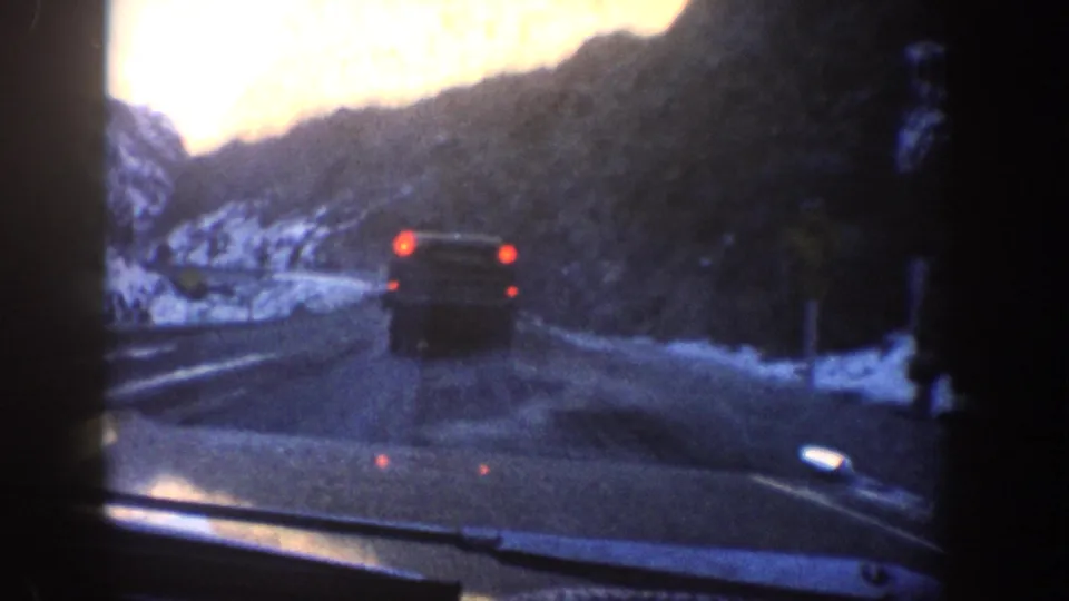 8mm windshield-POV shot following a car on a snow-lined mountain road with rocky terrain and a roadside post, consistent with Colorado mountain driving circa 1961.