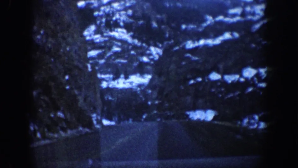 8mm home-movie frame showing a steep snow-covered cliff face from a moving vehicle, likely in the Colorado Rockies circa 1961, with a blue-toned color cast and visible film grain.