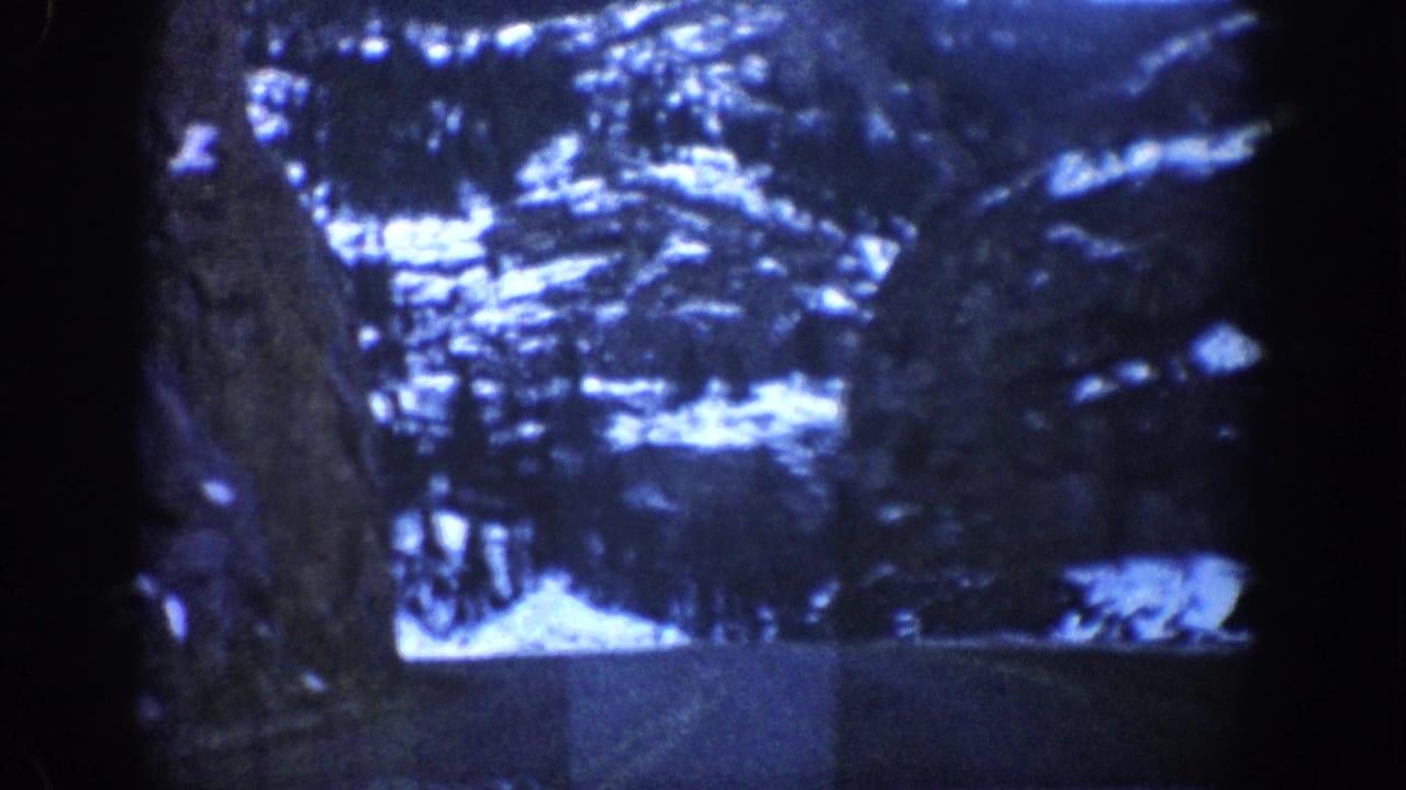 Dark blue-tinted 8mm frame showing a rocky canyon wall and snow-covered evergreen trees alongside a mountain road.