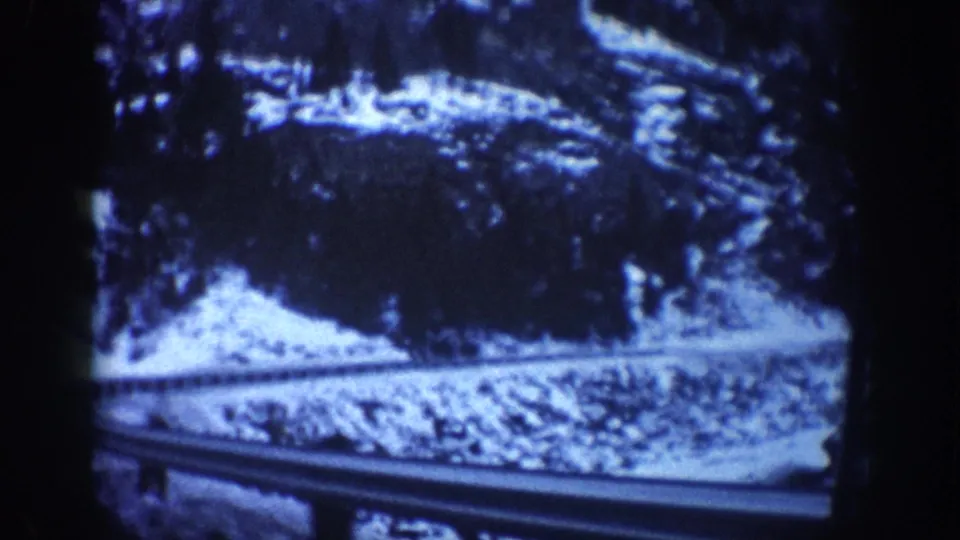 Passenger-perspective 8mm shot of a snow-banked Colorado mountain road with metal guardrail and conifer slopes, circa 1961.