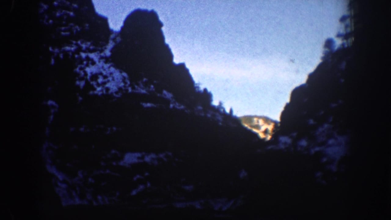 8mm still from a 1961 Aspen, Colorado winter drive; snow-dusted cliffs and winter sky visible through windshield, lower portion near-black.