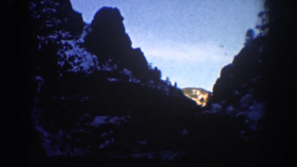 8mm still from a 1961 Aspen, Colorado winter drive; snow-dusted cliffs and winter sky visible through windshield, lower portion near-black.