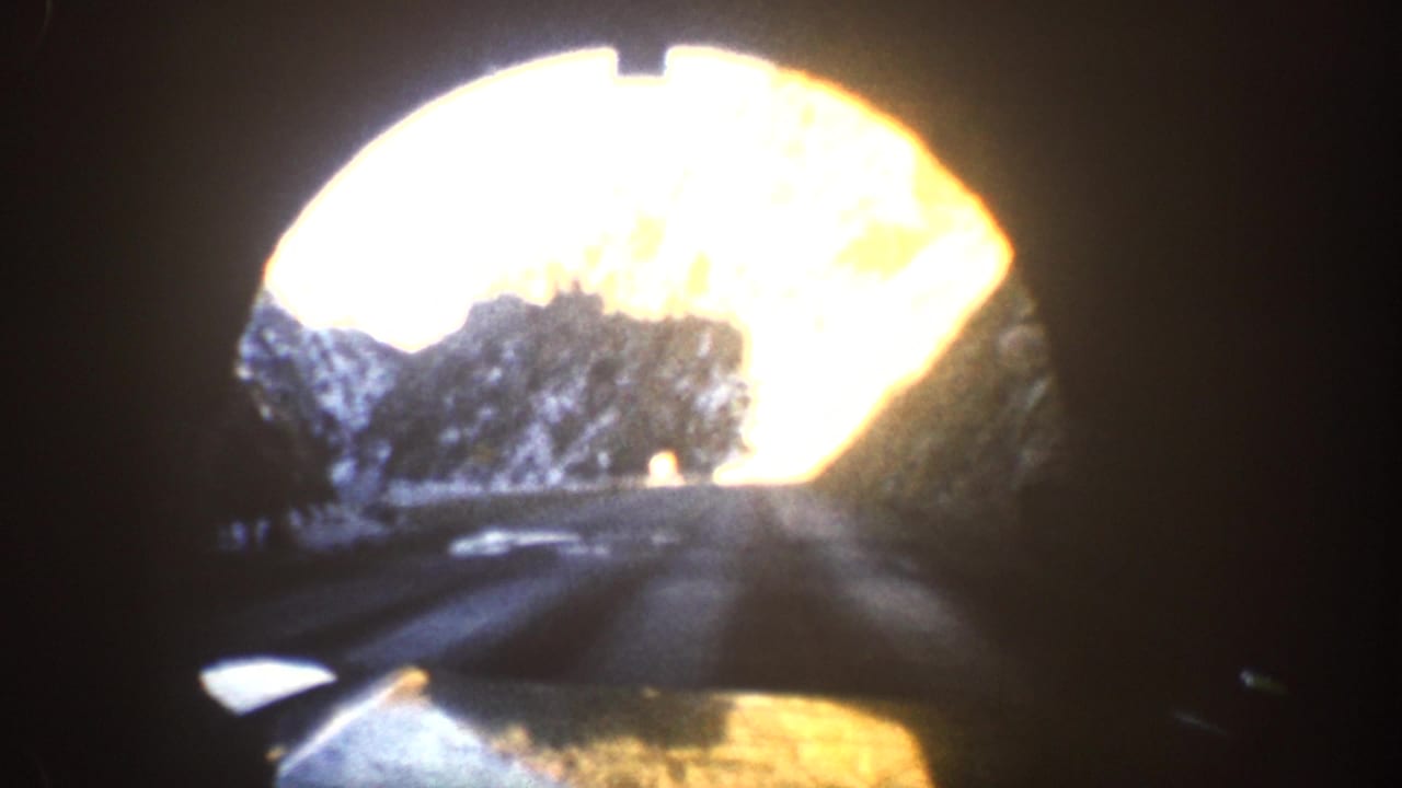 Dark vignette view through a circular tunnel opening onto a mountain road, car hood visible at the bottom of frame.