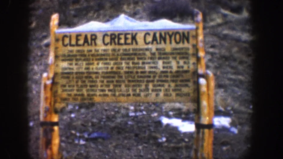 8mm second frame of the 'Clear Creek Canyon' wooden roadside interpretive sign on timber posts, snow patches on rocky ground, Colorado circa 1961.