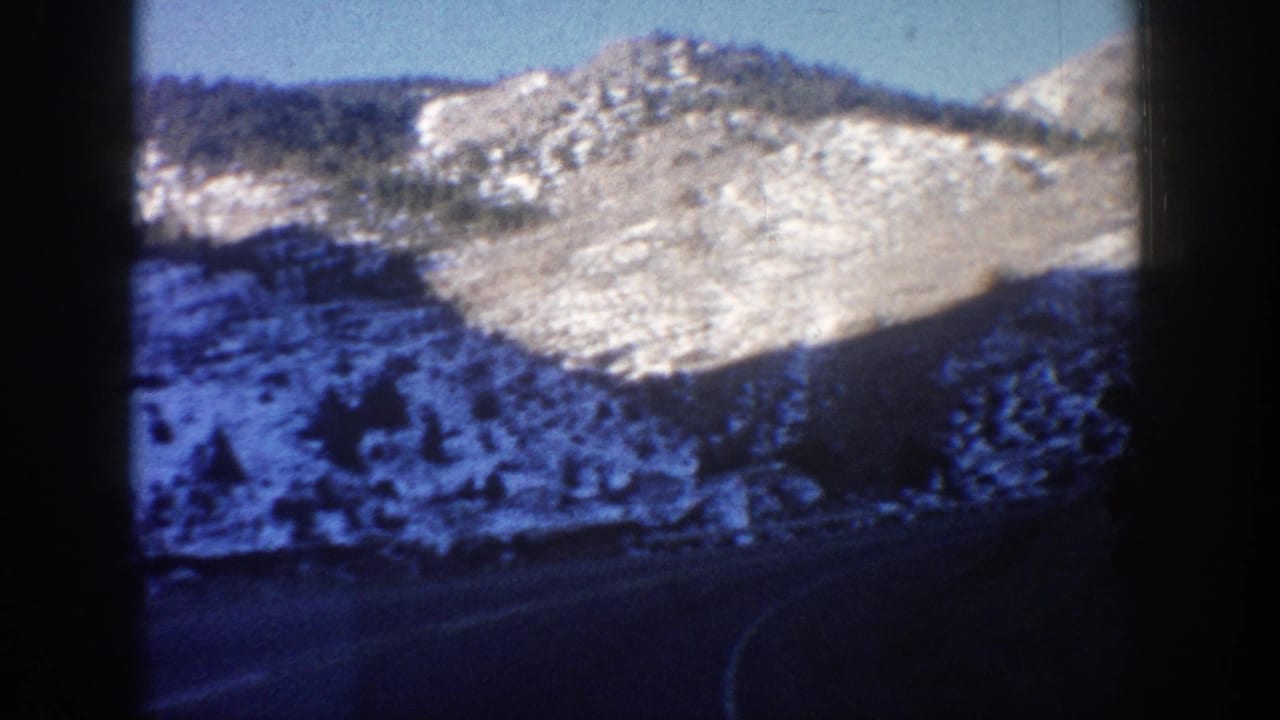 8mm home movie frame from a Colorado mountain road drive, circa 1961; snow-dusted rocky ridge and sparse conifers seen through a car window.