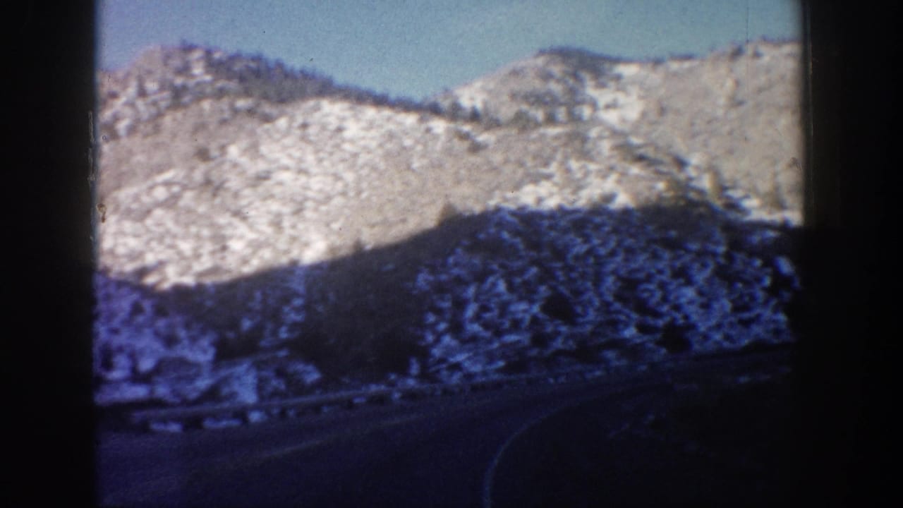 Rocky mountain ridge with light snow dusting, sparse conifers, and a curving road below, shot through a car window on 8mm film.