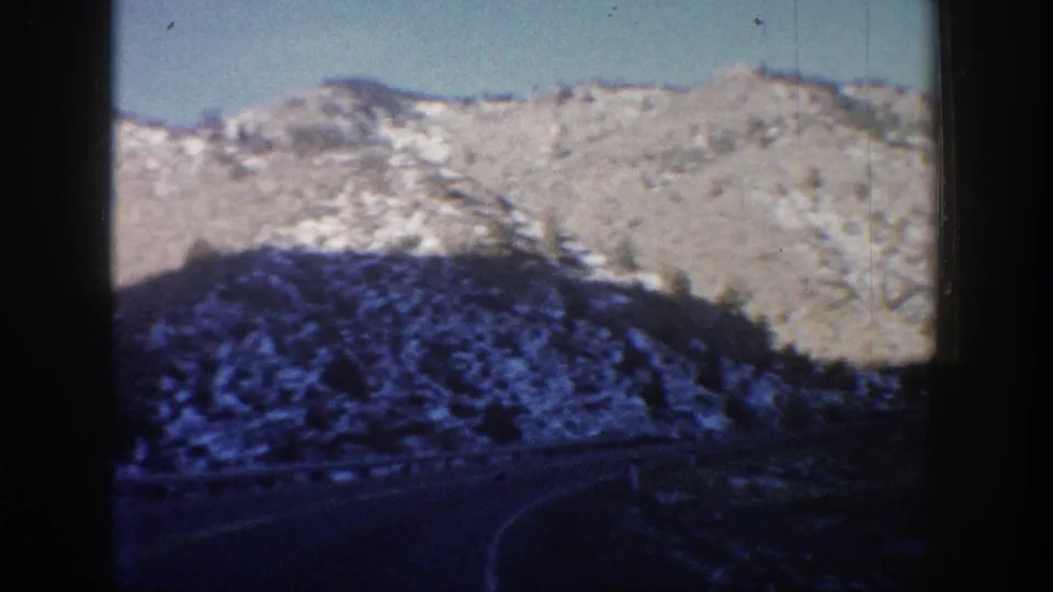 8mm home movie frame showing a rocky Colorado mountain slope with scattered evergreens and a road, viewed from a moving car, circa 1961.