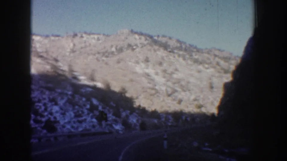 8mm frame from a Colorado mountain road drive circa 1961; rocky ridge, sparse conifers, and a dark foreground silhouette at frame right.