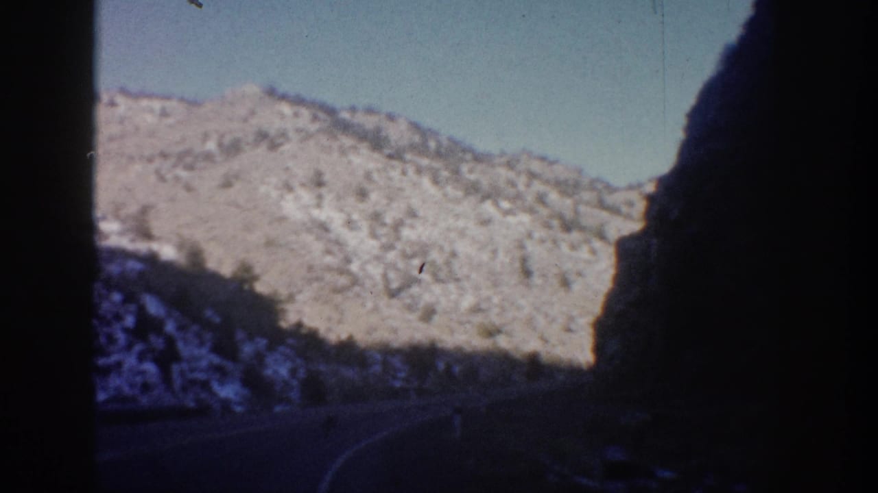 1961 Colorado road-trip view from inside a moving vehicle, showing a shaded road corridor, snow-dusted mountains, and pale blue sky.