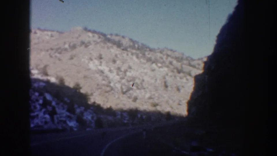 1961 Colorado road-trip view from inside a moving vehicle, showing a shaded road corridor, snow-dusted mountains, and pale blue sky.