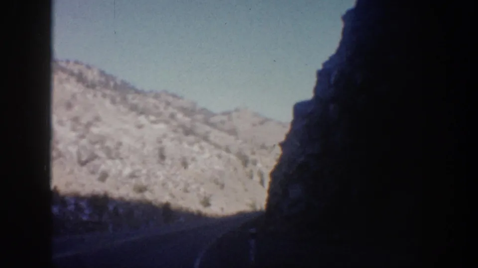 Passenger-window view of a curving Colorado mountain road with deep canyon shadow, sunlit snowy slopes, and blue sky.
