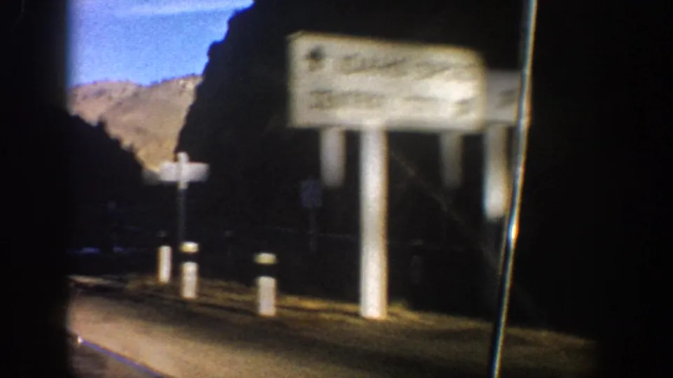 1961 Colorado road-trip frame with roadside signs, posts, a mountain backdrop, and heavy shadow from the vehicle-window view.