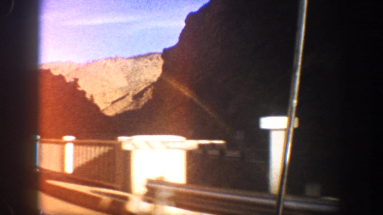 Moving car view of a white bridge railing, dark canyon walls, and sunlit mountains beyond.