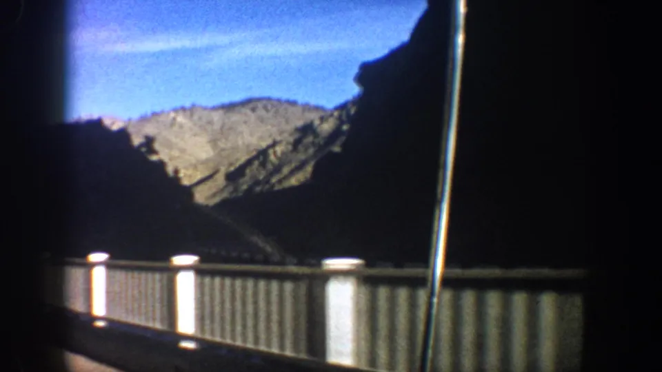 Vehicle-window view in the Aspen area, 1961, with a bridge railing sliding past dark mountain slopes and blue sky.