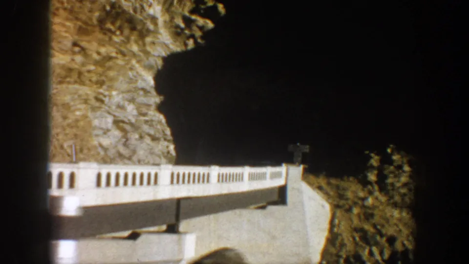 1961 8mm road-travel frame showing a white-railed bridge or roadside barrier beside a steep rocky canyon wall near Aspen.