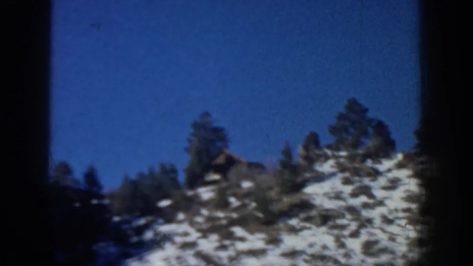 Aspen-area 1961 home-movie frame of a rocky hillside with patches of snow and pine trees under a saturated blue sky.