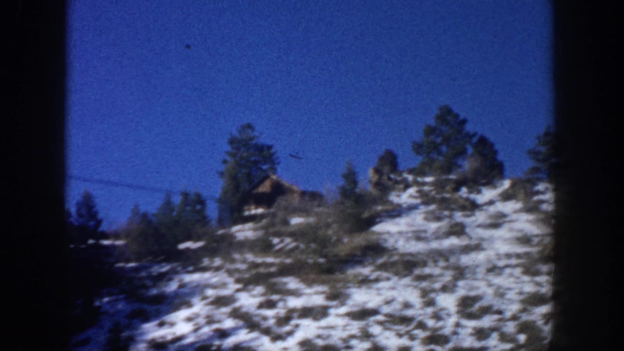 8mm home movie frame, Aspen Colorado area c. 1961 — snowy hillside, evergreen trees, and a hillside cabin visible through a car windshield.