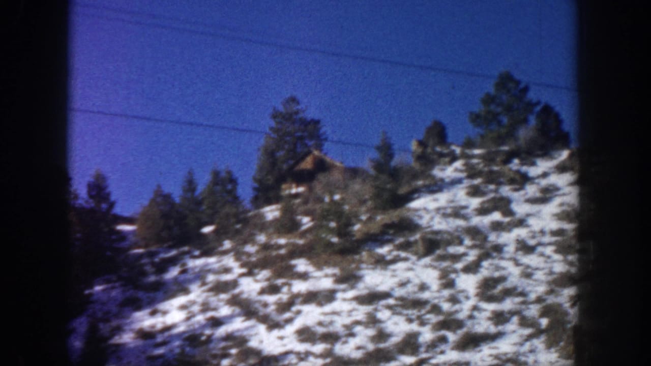 8mm film frame showing a snowy Rocky Mountain hillside with cabin and pines through a car windshield, Aspen Colorado area c. 1961.