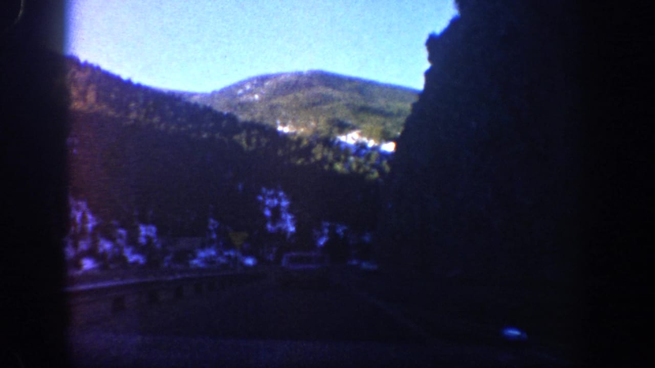 1961 Aspen, Colorado 8mm frame showing a road, forested mountain slopes, snow patches, and bright sky through a dark vehicle interior.