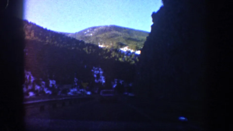 1961 Aspen, Colorado 8mm frame showing a road, forested mountain slopes, snow patches, and bright sky through a dark vehicle interior.