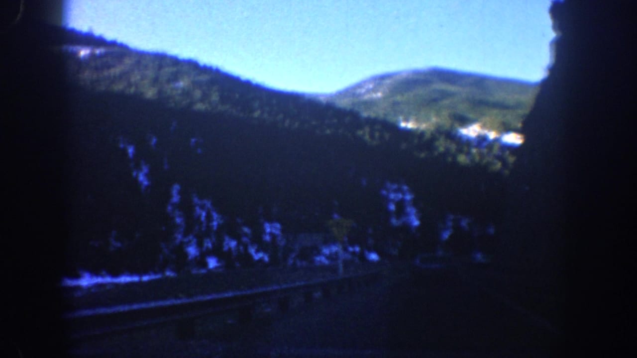 1961 Aspen, Colorado travel footage frame with evergreen-covered hills, a road edge, snow patches, and heavy shadow from the car interior.