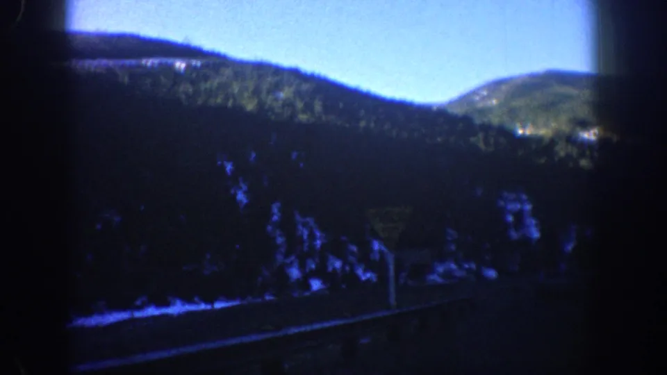 1961 Aspen, Colorado 8mm frame showing a roadside forest, rounded mountain ridge, blue sky, and dark vehicle-window framing.