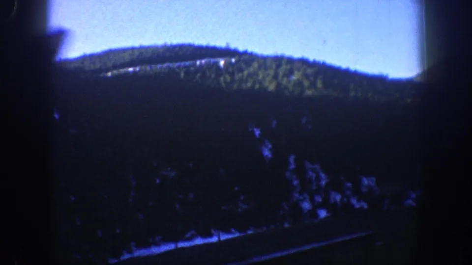 1961 Aspen, Colorado frame with evergreen hillside, road line, snow traces, and bright sky partly obscured by the vehicle interior.