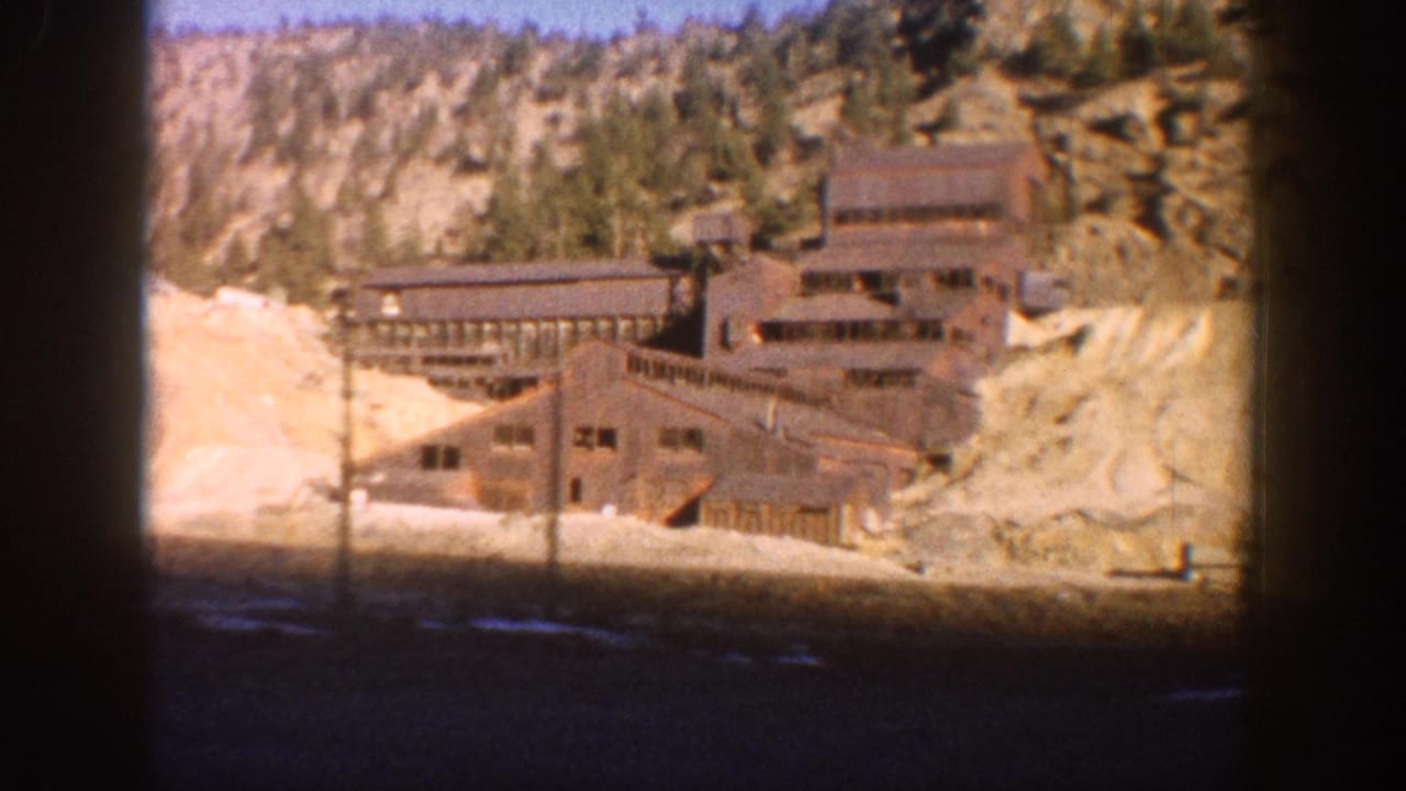 Multi-story weathered wooden mining mill building against a rocky hillside with evergreen trees, dirt foreground, and utility poles.