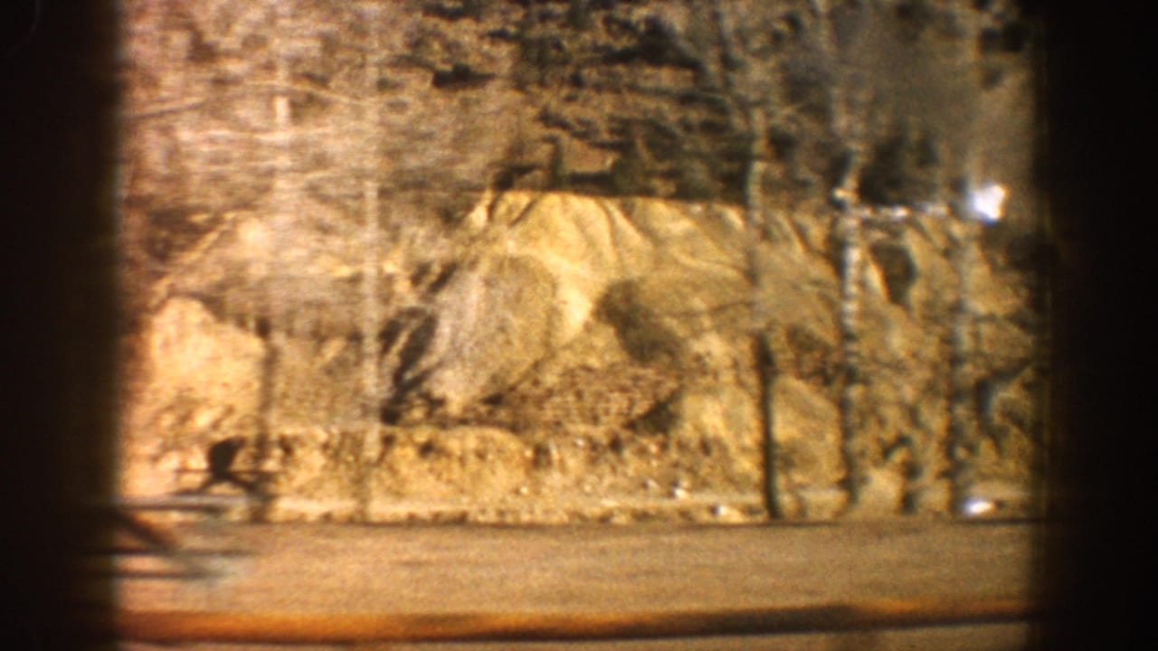 Close pass of an exposed rocky cliff or cut bank, ochre and tan tones, filmed from a moving vehicle with viewfinder vignette.