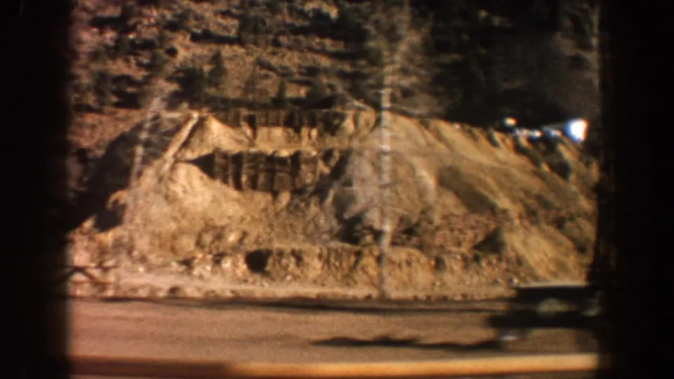 Earthen spoil or tailings mound alongside the road near Aspen, Colorado, 1961, with a conifer-covered slope above, filmed on 8mm.