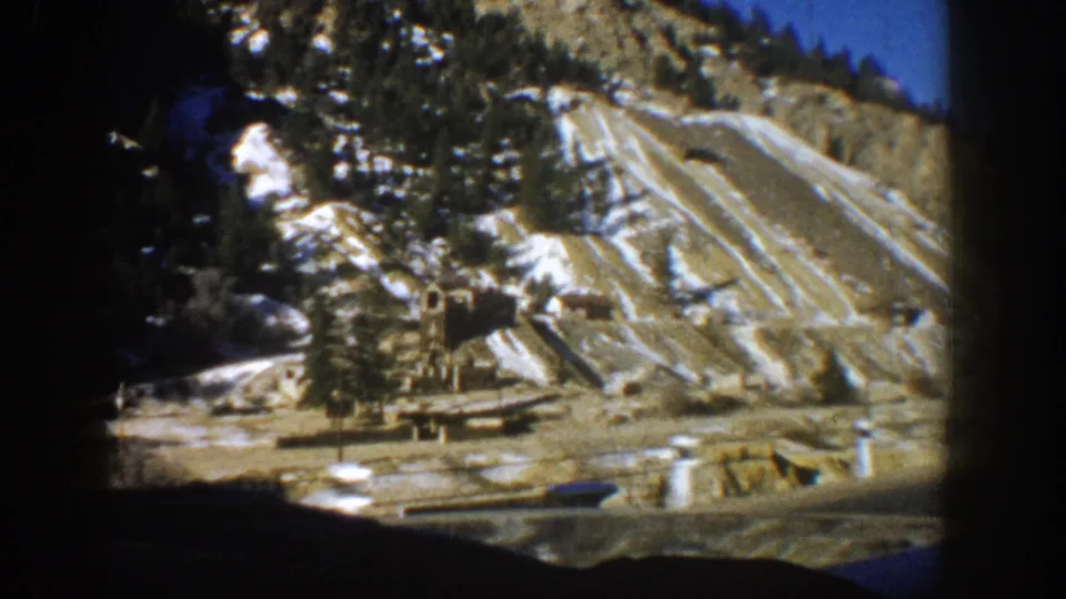 8mm frame showing layered rock strata with snow, evergreen trees, and ground-level industrial equipment at a Colorado mine site exterior, circa 1961.