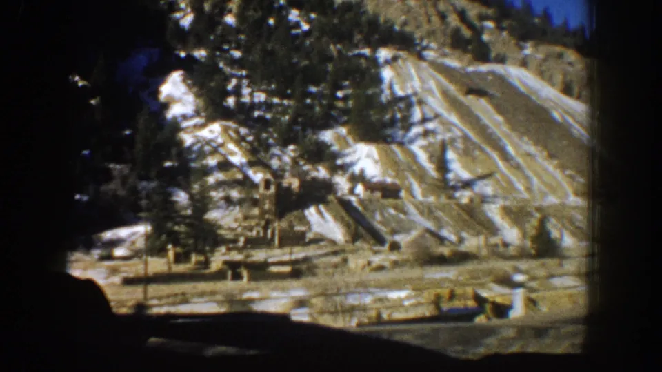 8mm frame of a stratified snow-patched hillside with evergreen trees and scattered ground equipment at a Colorado mine exterior, circa 1961.